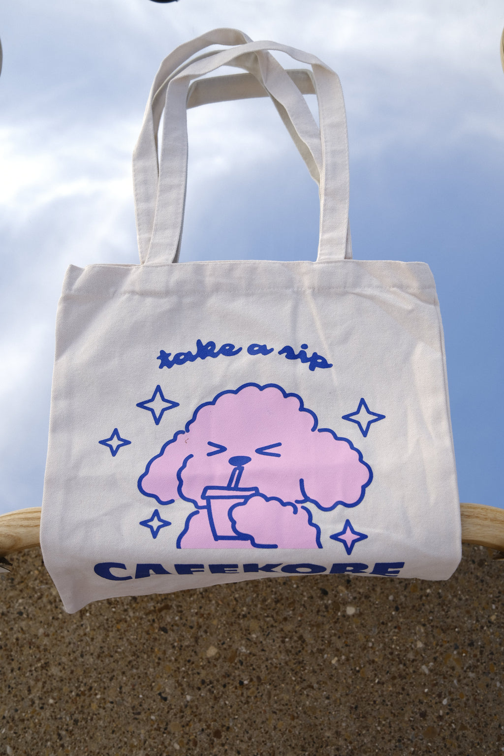 White tote bag with pink cloud design and blue text against a clear sky.