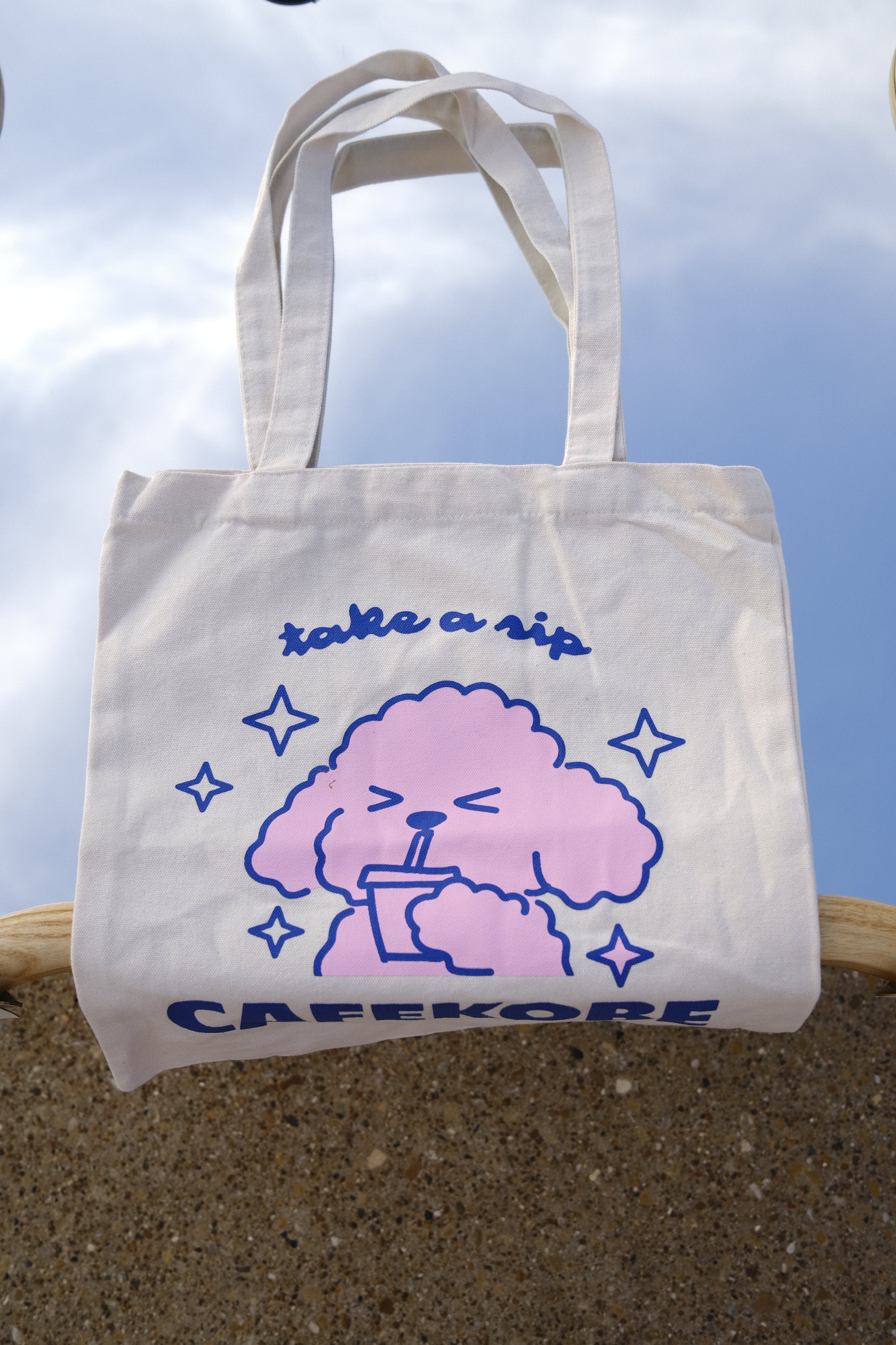 White tote bag with pink cloud design and blue text against a clear sky.